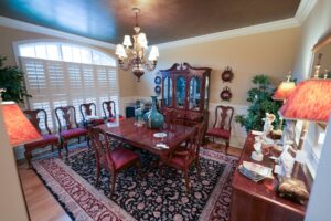 A dining room with a wooden table and chairs, chandelier above, and a china cabinet against the wall. The room is decorated with plants, matching lamps, a patterned rug, and a large window with shutters.
