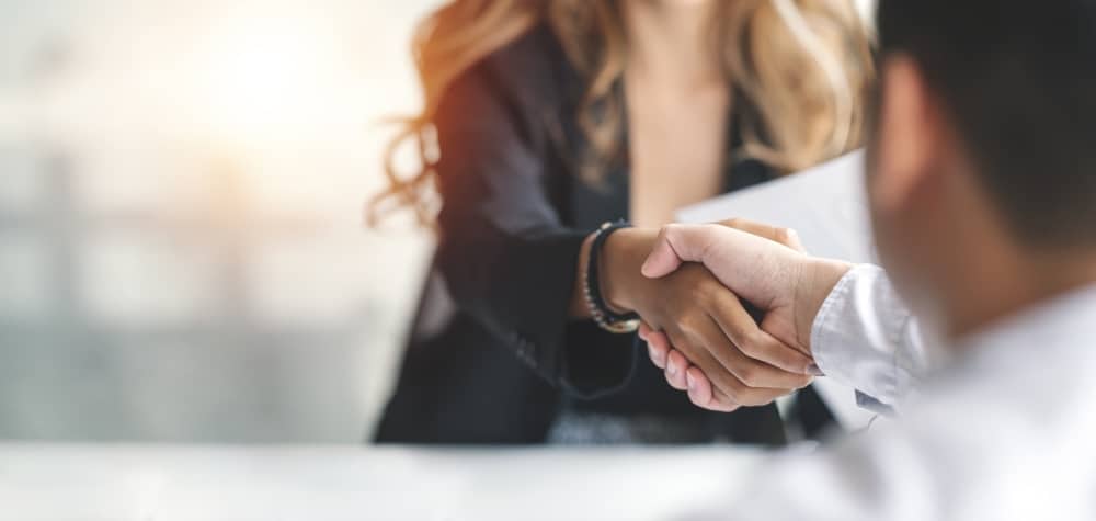 Two people are shaking hands across a table, suggesting an agreement or partnership. The focus is on their hands, with one person wearing a dark outfit and the other in white. The background is softly blurred, creating a professional and neutral setting.