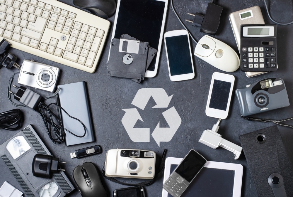 A collection of outdated electronic devices, including phones, cameras, a keyboard, and a calculator, surrounds a recycling symbol on a dark surface, symbolizing e-waste recycling.