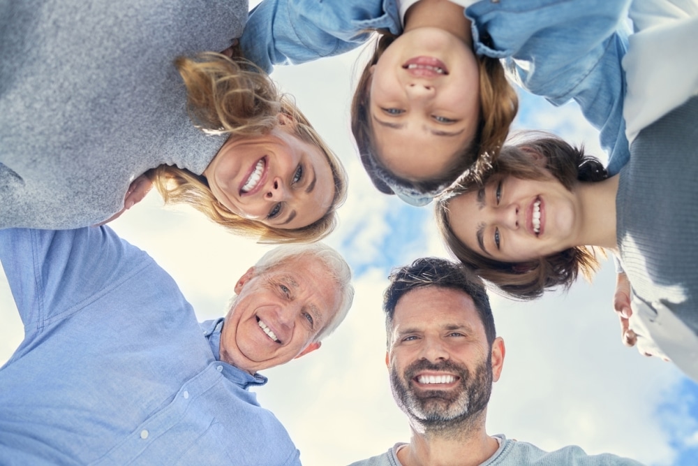 A group of five people, including two adults and three children, are huddled in a circle, smiling and looking down at the camera against a blue sky background.