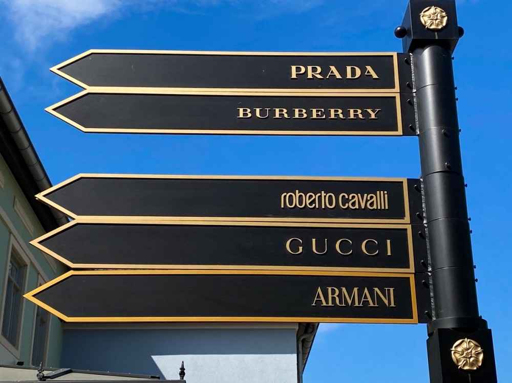 A street sign with five directional arrows pointing to Prada, Burberry, Roberto Cavalli, Gucci, and Armani against a blue sky background.