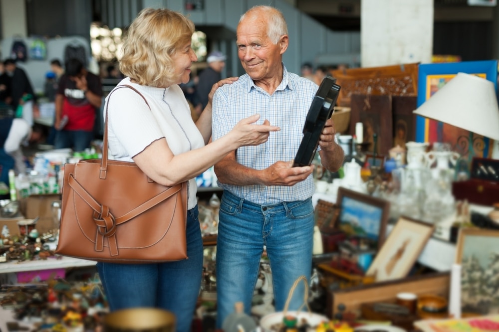 An elderly couple examines a black bottle at an estate sale. The woman, holding a brown purse, gestures while the man in a checkered shirt and jeans holds the bottle. Various items are displayed around them on a table.