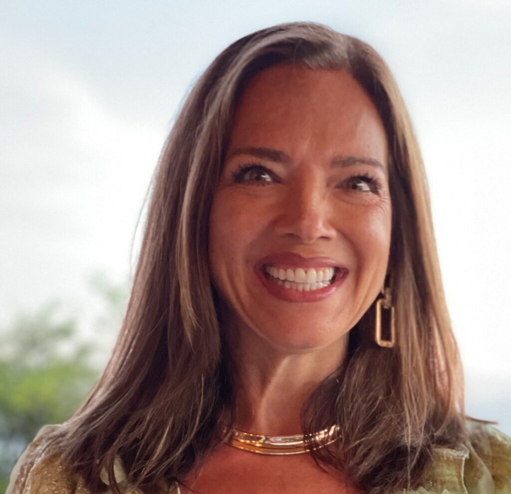 A woman with long brown hair smiles brightly. She is wearing a light-colored top, a gold necklace, and hoop earrings. The background is softly blurred, suggesting an outdoor setting.