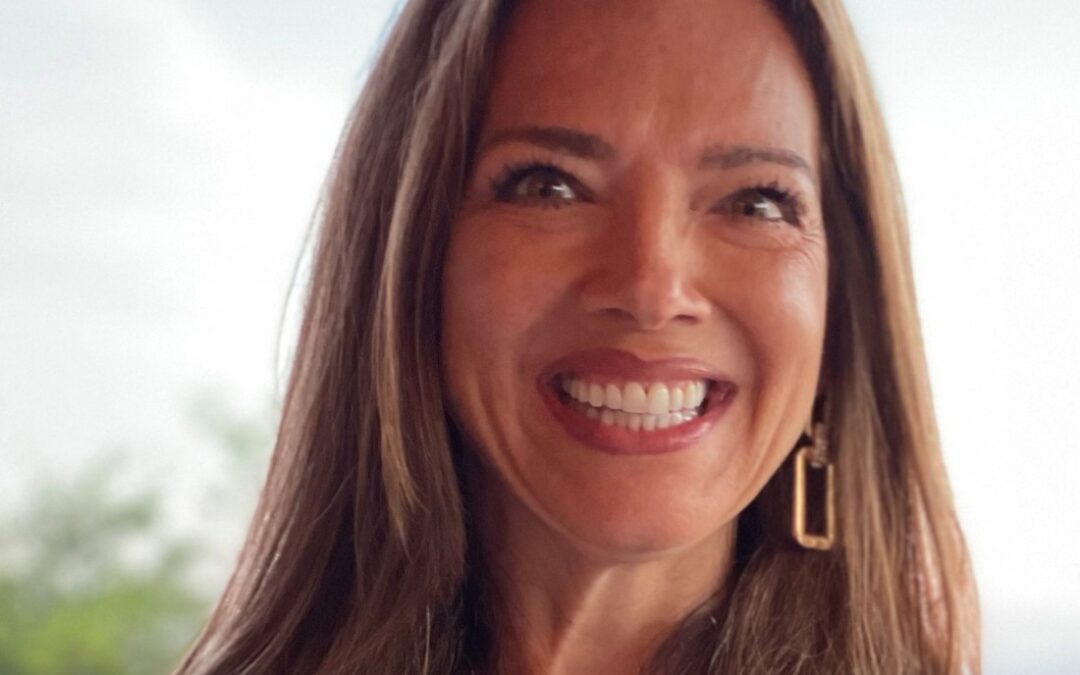 A woman with long brown hair smiles brightly. She is wearing a light-colored top, a gold necklace, and hoop earrings. The background is softly blurred, suggesting an outdoor setting.