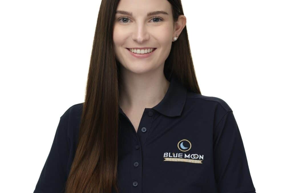 A person with long brown hair is smiling and wearing a navy blue polo shirt with a "Blue Moon" logo on the chest. The background is white.