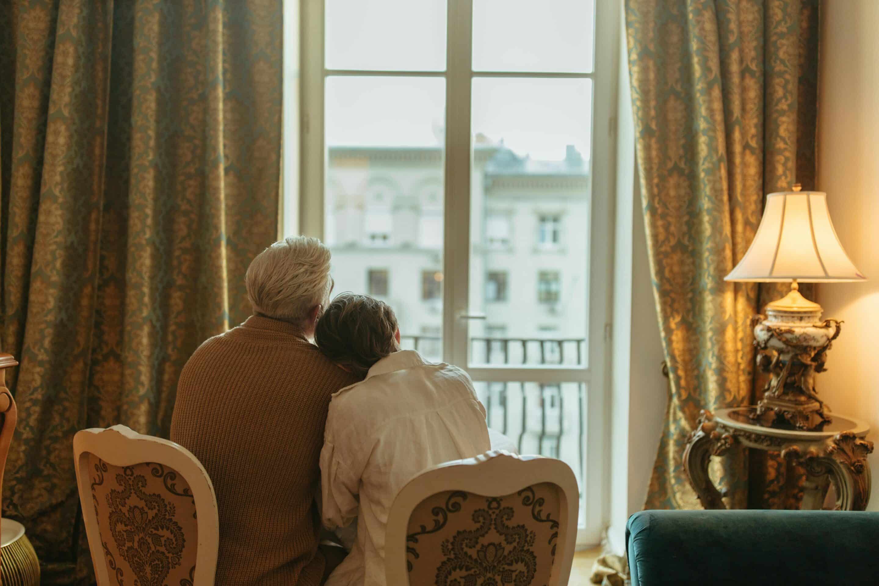 A husband and wife comfort each other while looking out the windows of their beautifully decorated home.