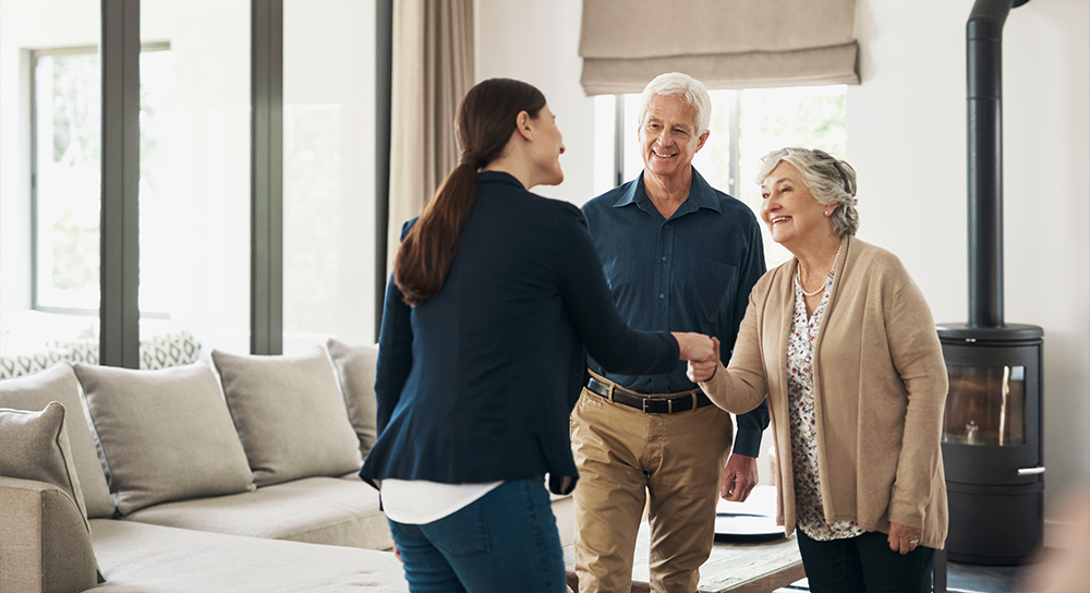A woman shakes hands with an elderly couple to welcome them into the eldercare franchise space.
